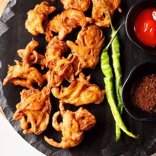 onion pakoda on a a black granite board with fried green chillies and condiments in two black bowls.