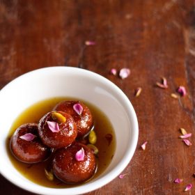 gulab jamun with milk powder garnished with dried rose petals and pistachios and served in a white bowl.