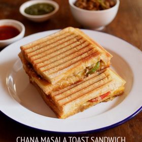 chana sandwich placed on white plate with a white bowl of chana masala, and two small white bowls of green chutney and tomato ketchup.
