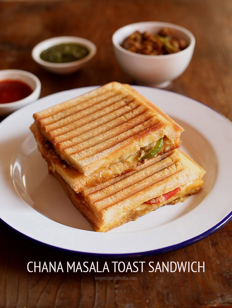 chana sandwich placed on white plate with a white bowl of chana masala, and two small white bowls of green chutney and tomato ketchup.