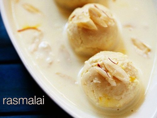 4 balls of rasmalai in an oblong white serving dish covered with thickened milk and garnished with sliced almonds and saffron threads