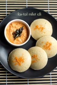 millet idli sprinkled with podi and served on a black plate with a side bowl of coconut chutney.