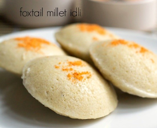 millet idli sprinkled with podi and served on a white plate with a side of bowls of sambhar and coconut chutney.