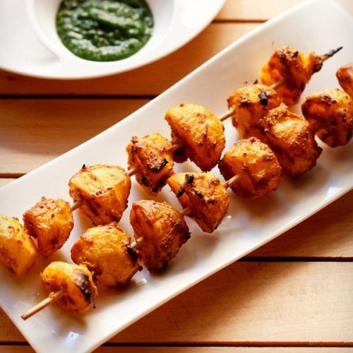 tandoori aloo tikka served on a white rectangular platter with a bowl of green chutney on the top left side and text layovers.