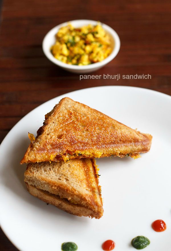 paneer bhurji sandwich pieces serve don a white plate with green chutney and tomato ketchup.