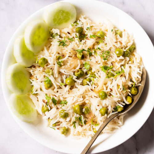 overhead shot of green peas pulao in a white shallow bowl with a brass spoon and cucumber slices placed at the left side on a white marble table