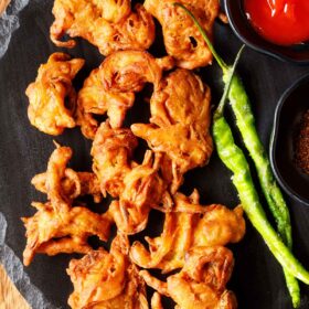 onion pakoda on a black round platter served with ketchup and fried green chillies.