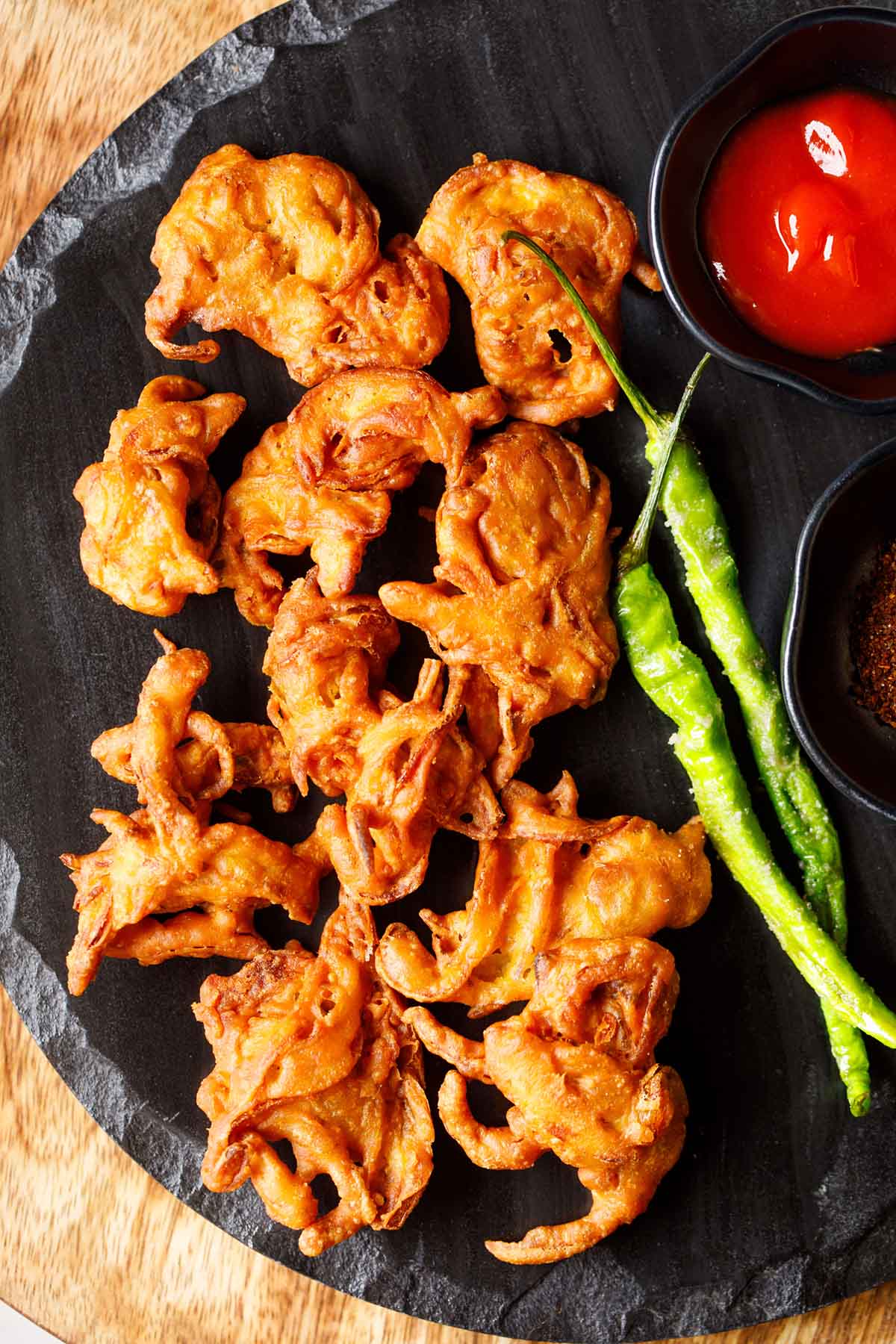 onion pakoda on a black round platter served with ketchup and fried green chillies.