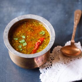 tomato rasam in a traditional South Indian container with a spoon placed on top of a white kitchen napkin on the left side on a dark navy blue board.