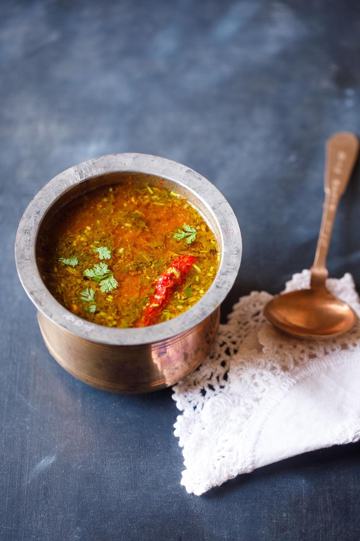 tomato rasam in a traditional South Indian container with a spoon placed on top of a white kitchen napkin on the left side on a dark navy blue board.