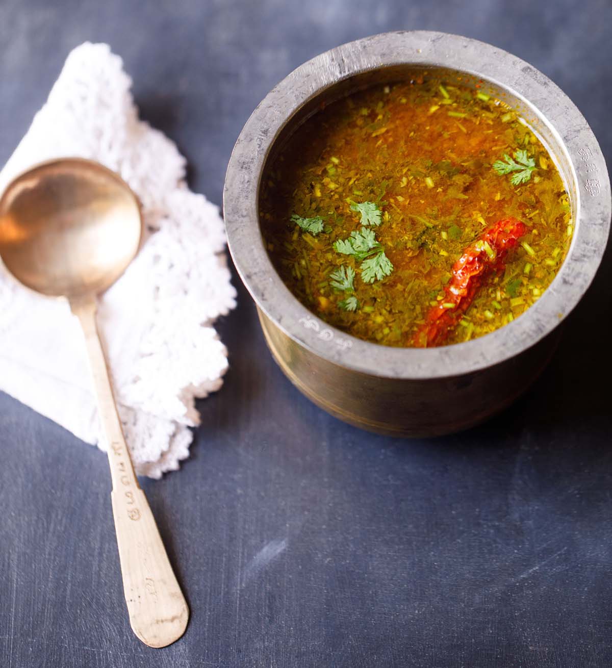 tomato rasam in a traditional South Indian container with a spoon placed on a white doily napkin next to the container on a dark blue board.