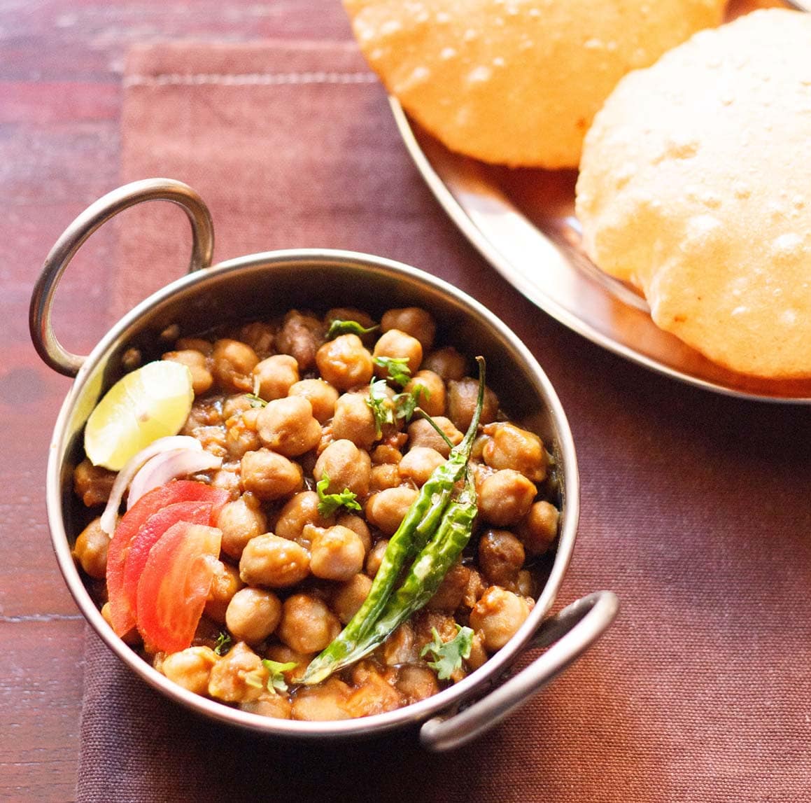 chana masala in a small serving wok topped with sliced tomatoes, onions, lemon and green chillies served with pooris.