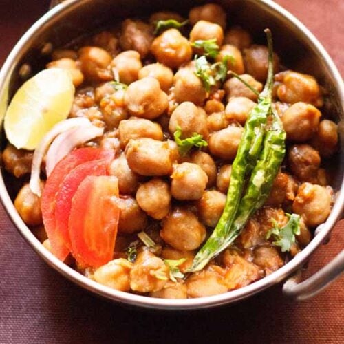 closeup image of chana masala in a small serving wok topped with sliced tomatoes, onions, lemon and green chillies.