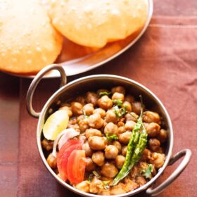 chana masala in a small serving wok topped with sliced tomatoes, onions, lemon and green chillies served with poori.