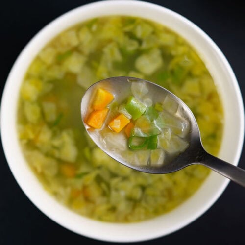 closeup image of clear soup with vegetables being seen in a steel spoon on top of white bowl with the soup on black table.