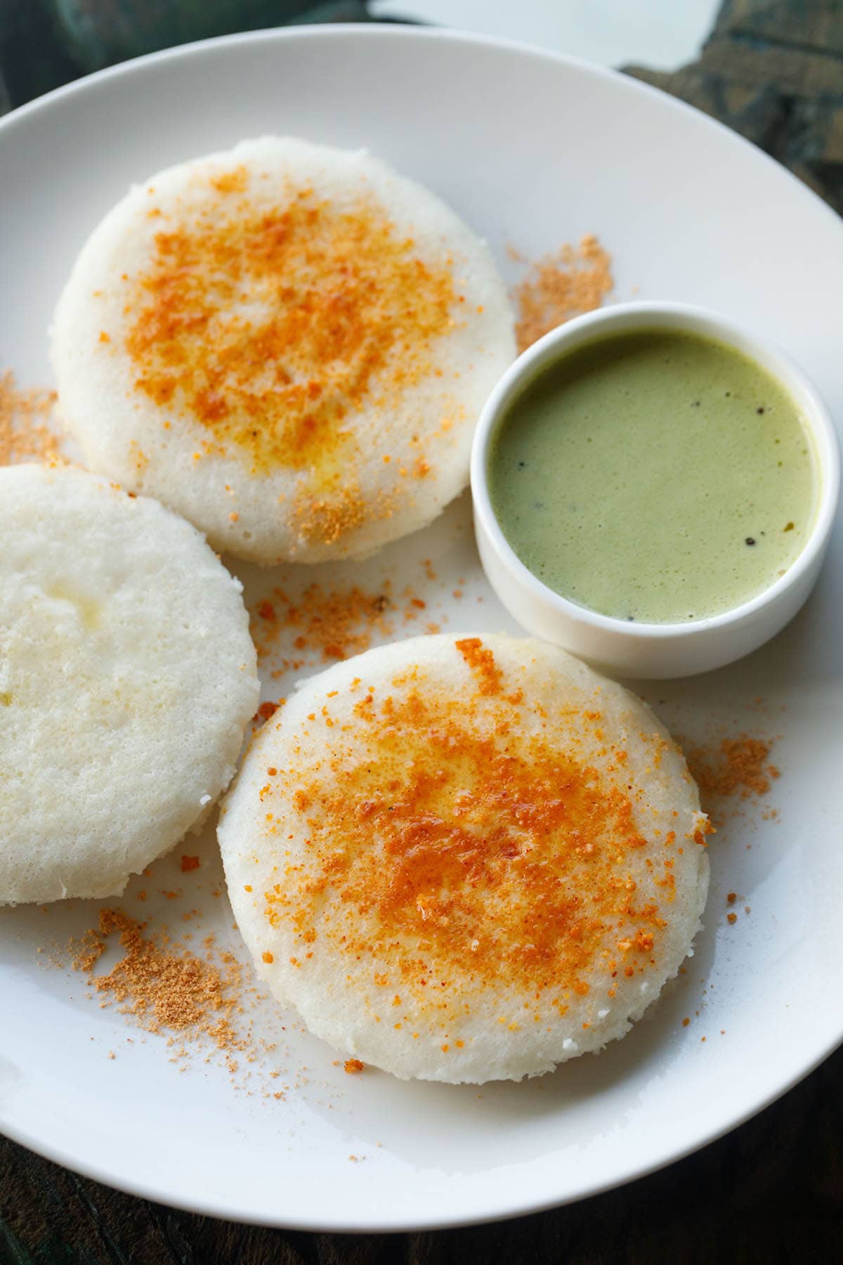 three plate idli sprinkled with podi and ghee on a plate with green coconut chutney by side. 