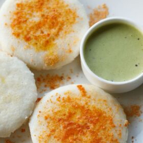 three thatte idli sprinkled with podi and ghee served on a plate with cilantro coconut chutney in a white bowl.