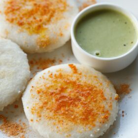 three thatte idli sprinkled with podi and ghee served on a plate with cilantro coconut chutney in a white bowl.