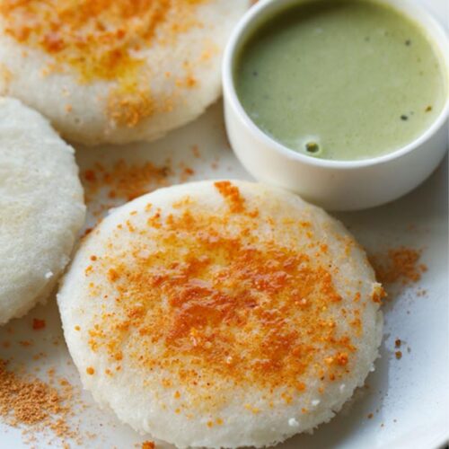 three thatte idli sprinkled with podi and ghee on a plate with green coconut chutney by side.