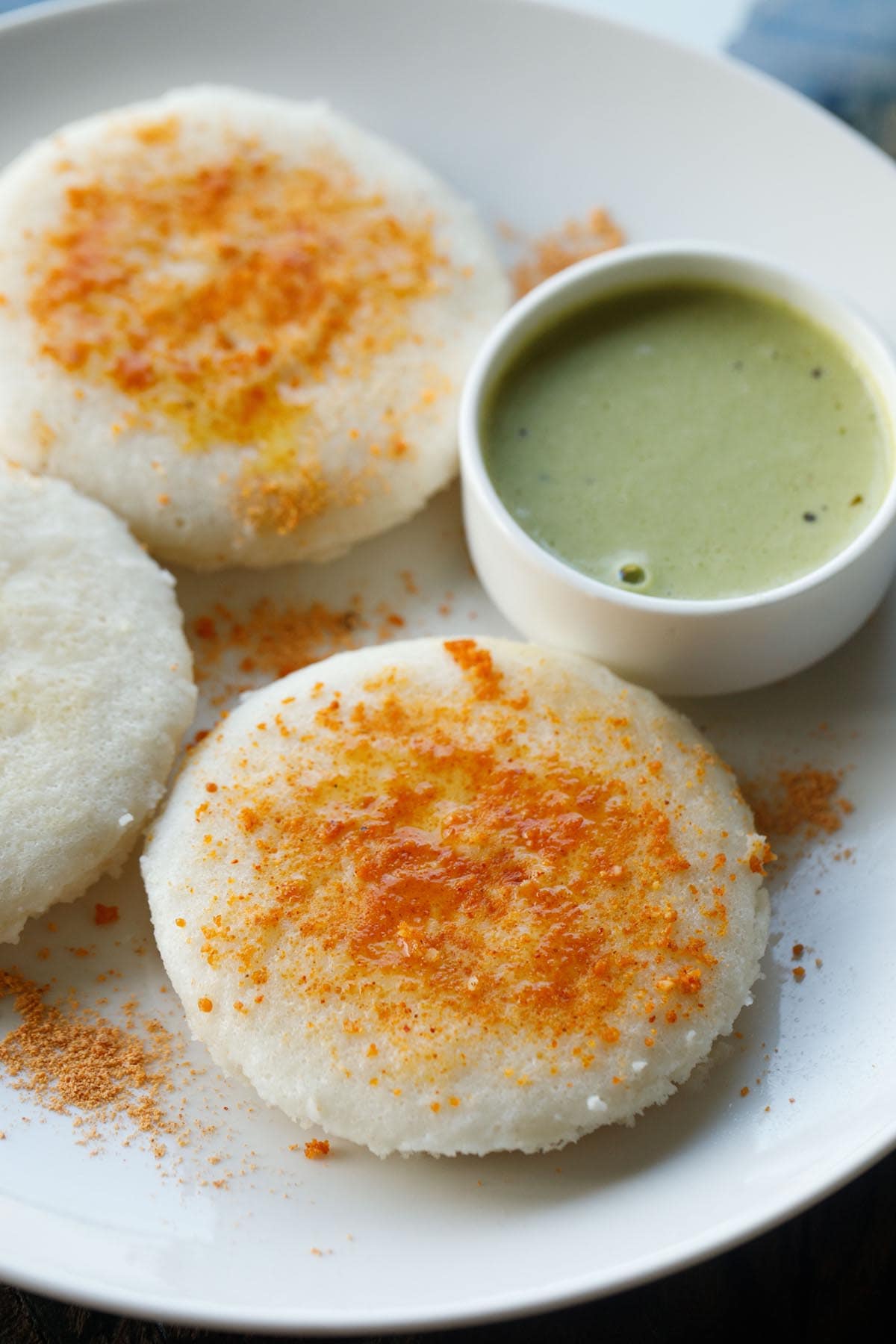 three thatte idli sprinkled with podi and ghee served on a plate with cilantro coconut chutney in a white bowl.