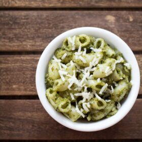 pesto pasta served in a white bowl topped with some grated parmesan on a dark brown wooden tray