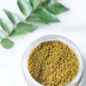 curry leaves powder in a small glass jar with a sprig of curry leaves above on a white background.