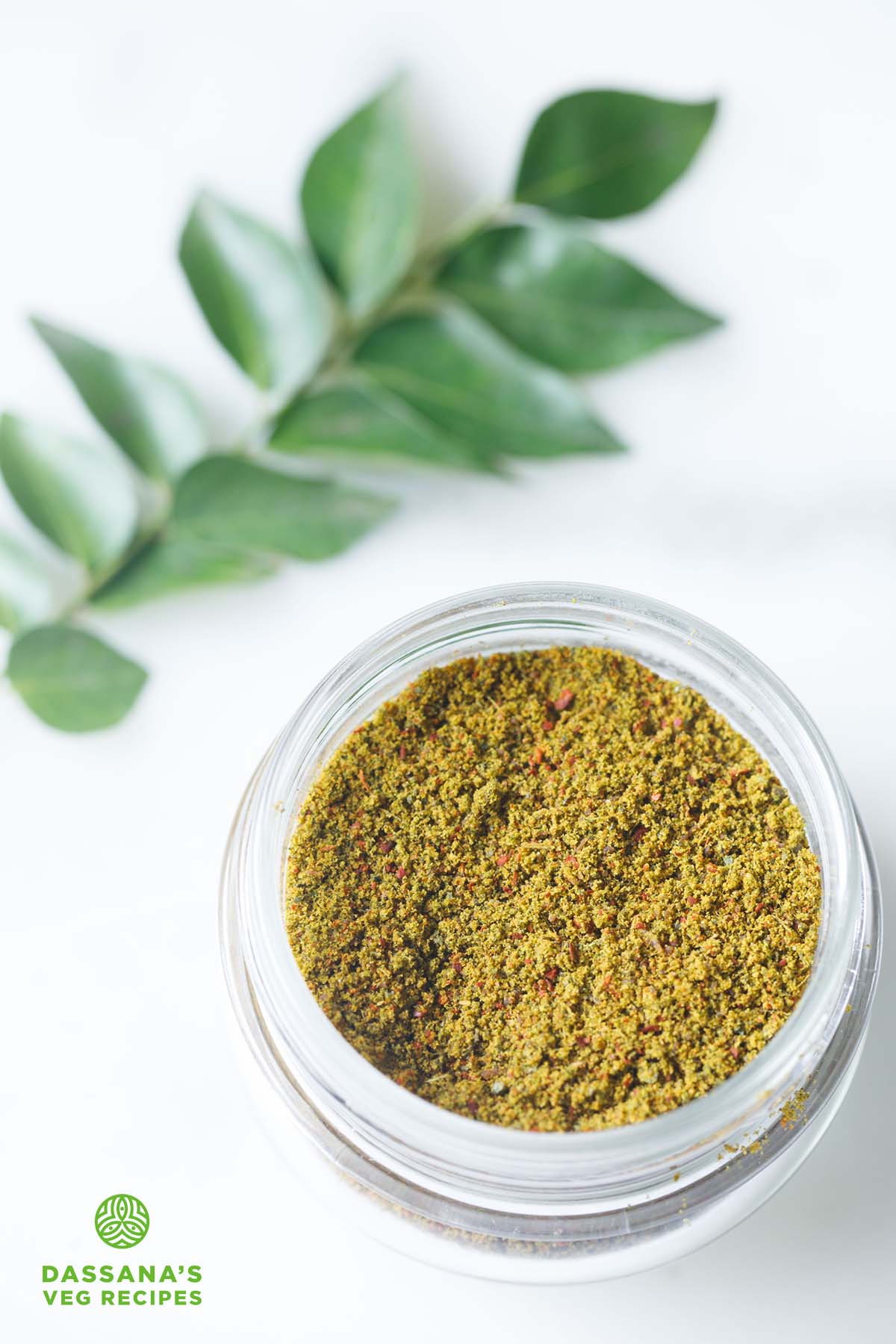 curry leaves powder in a small glass jar with a sprig of curry leaves above on a white background.