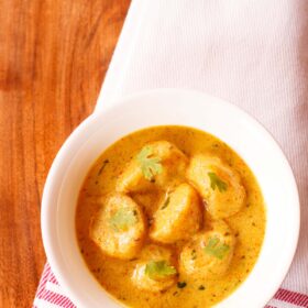 dum aloo garnished with coriander leaves and served in a white bowl on a red bordered white napkin.