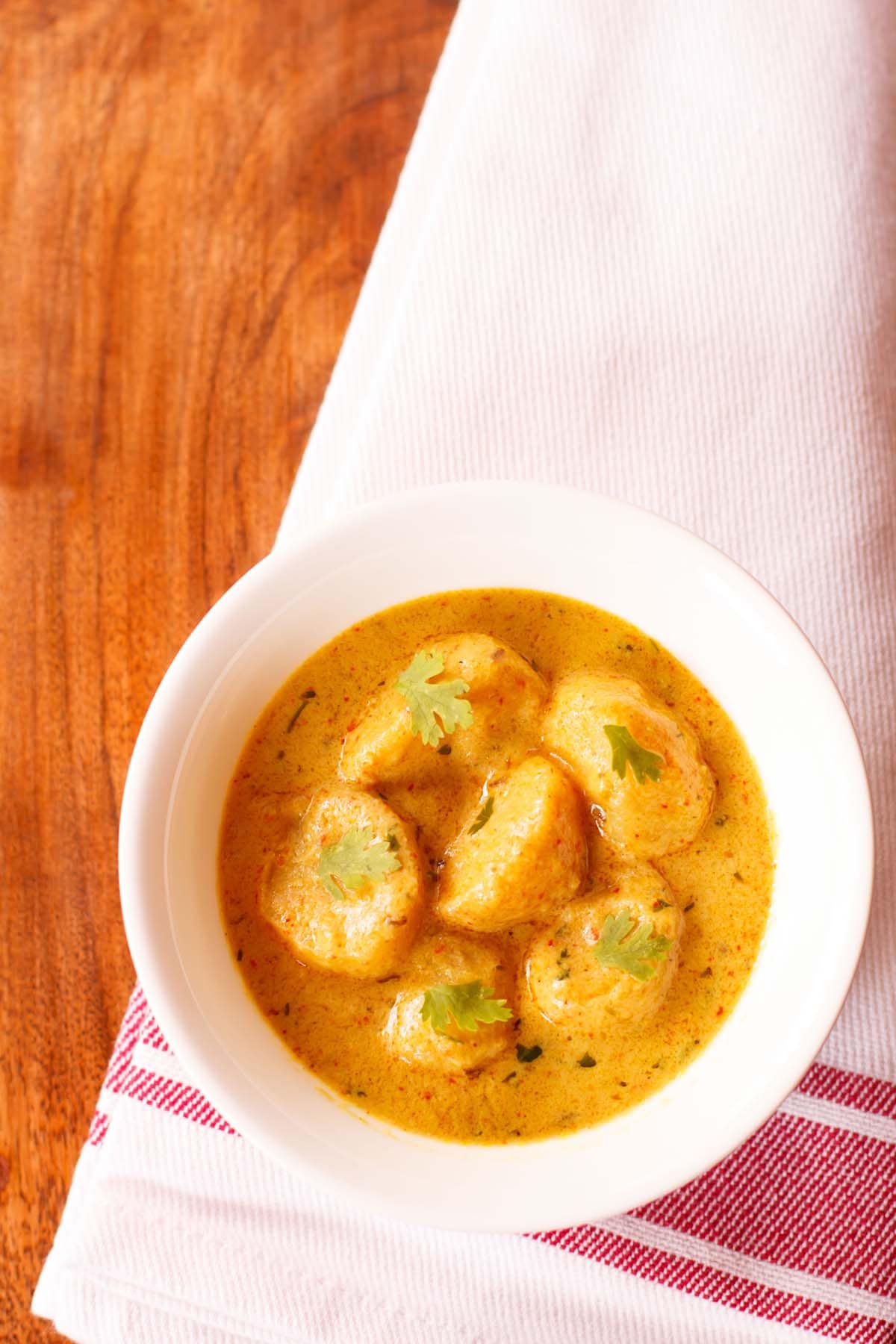 dum aloo garnished with coriander leaves and served in a white bowl on a red bordered white napkin.