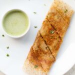 folded paneer dosa garnished with a few coriander leaves alongside a bowl of green colored coconut chutney.