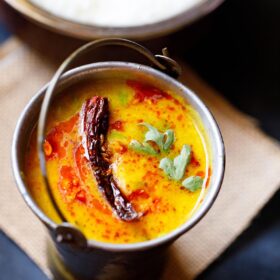 dal tadka garnished with cilantro and a topped with fried red chilli with some fried cumin and some red colored oil in a small brass bucket on a light brown jute mat with a bowl of steamed rice kept above on the top-right.