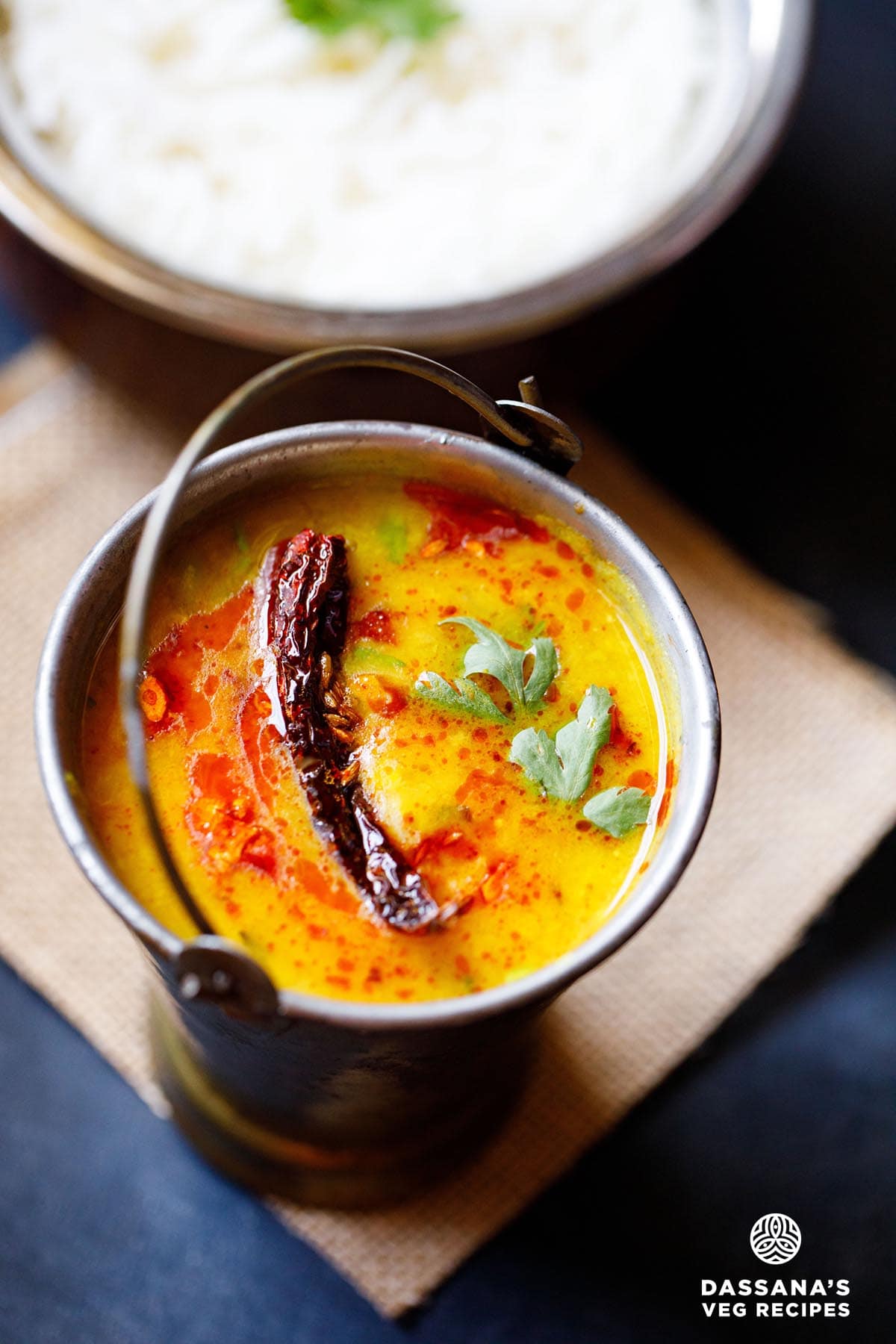 dal tadka garnished with cilantro and a topped with fried red chilli with some fried cumin and some red colored oil in a small brass bucket on a light brown jute mat with a bowl of steamed rice kept above on the top-right.