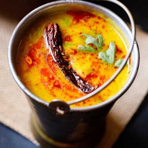 dal tadka garnished with cilantro and a topped with fried red chilli with some fried cumin and some red colored oil in a small brass bucket on a light brown jute mat.