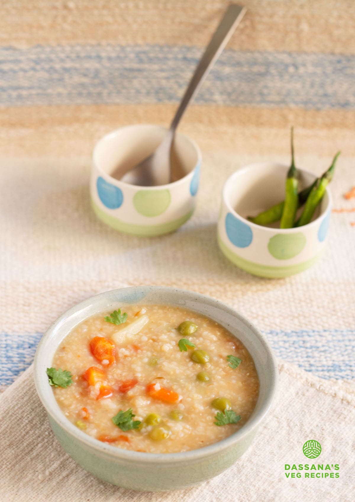 daliya khichdi served in a ceramic bowl with two small bowls on top, one with a spoon and the other with three green chillies on a jute fabric.