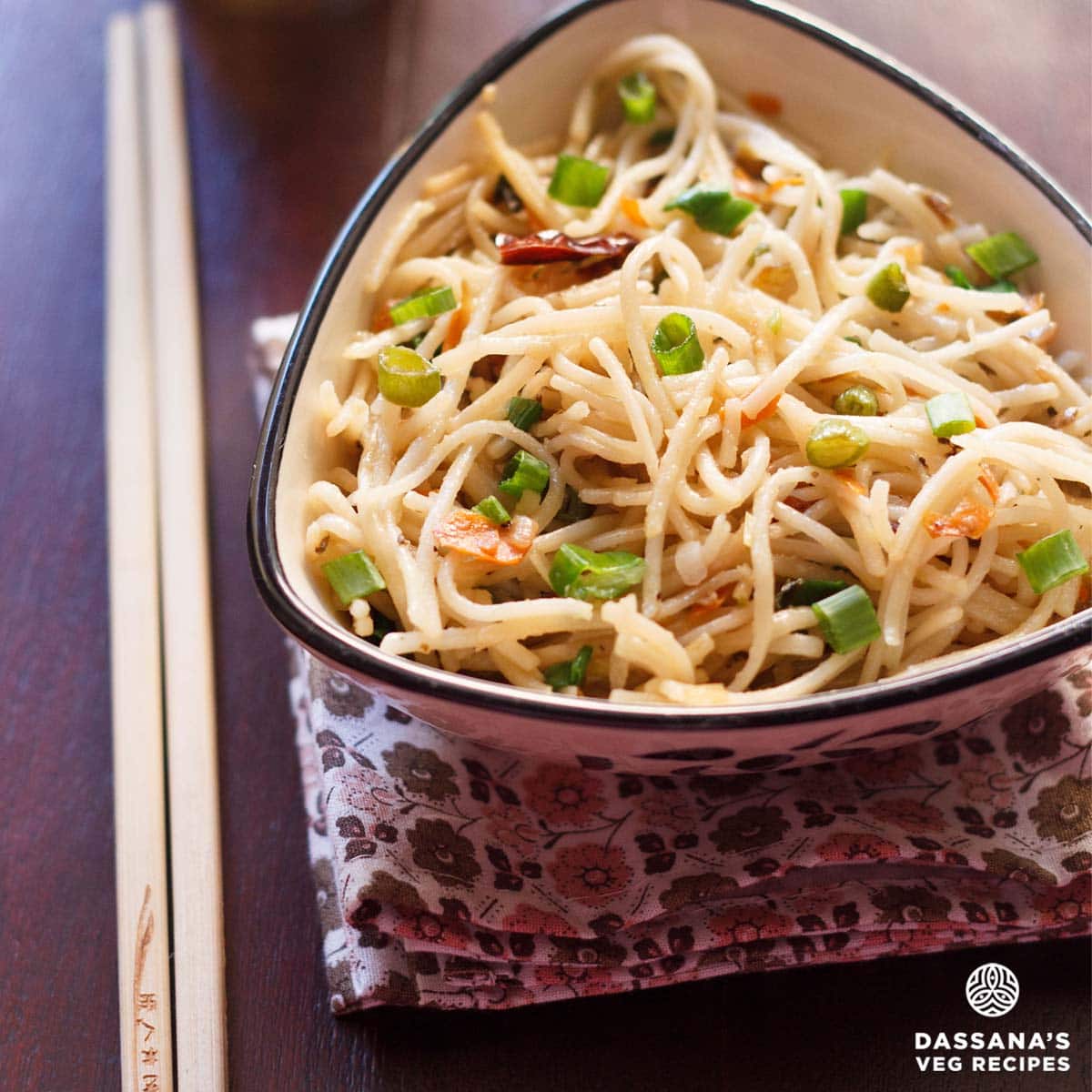 hakka noodles garnished with chopped spring onion greens and served in a triangular bowl on a printed napkin with cream colored bamboo chopsticks placed at the left side.