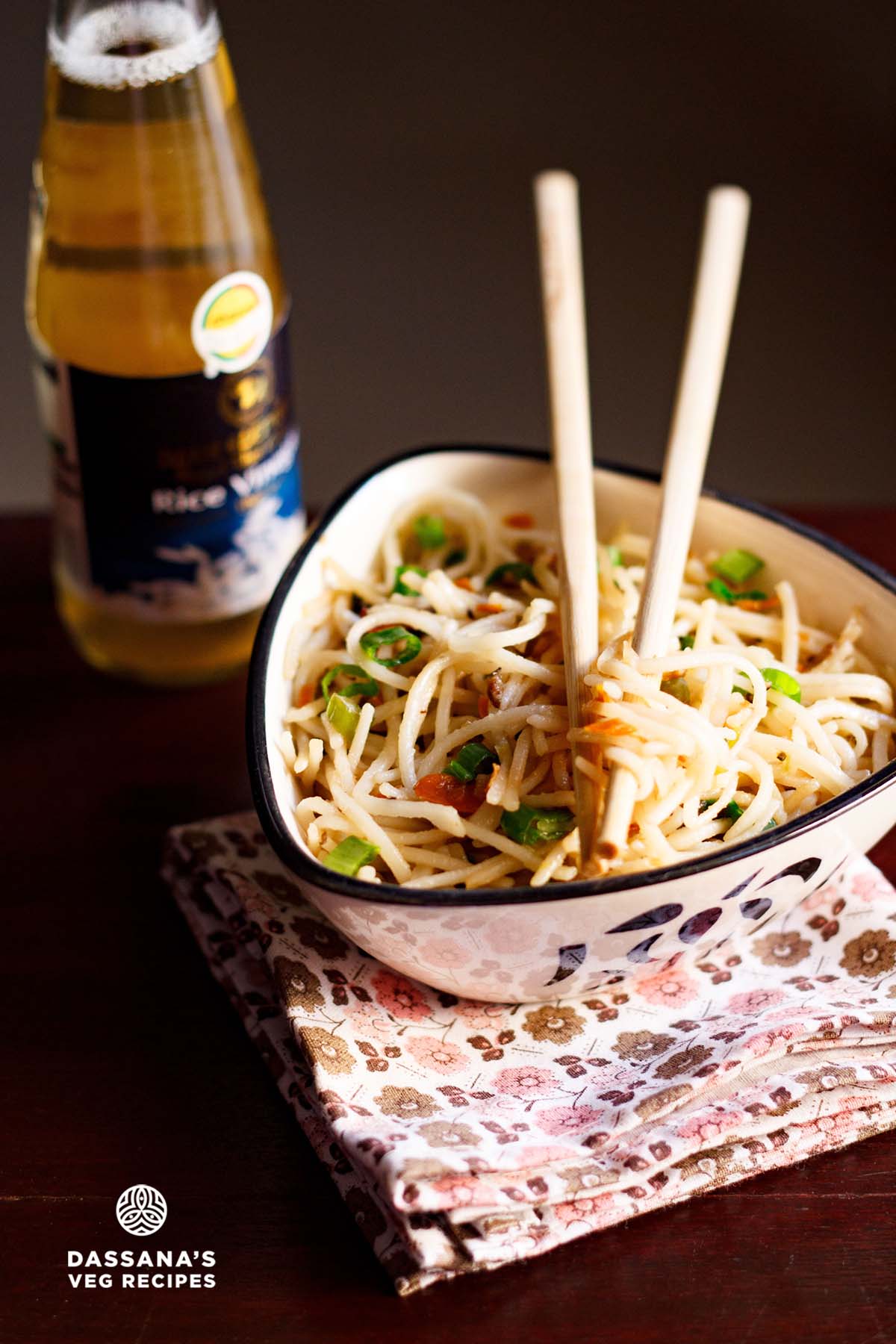 hakka noodles served in a triangular bowl on a printed napkin with cream colored bamboo chopsticks placed on top of the bowl with some hakka noodles between them.