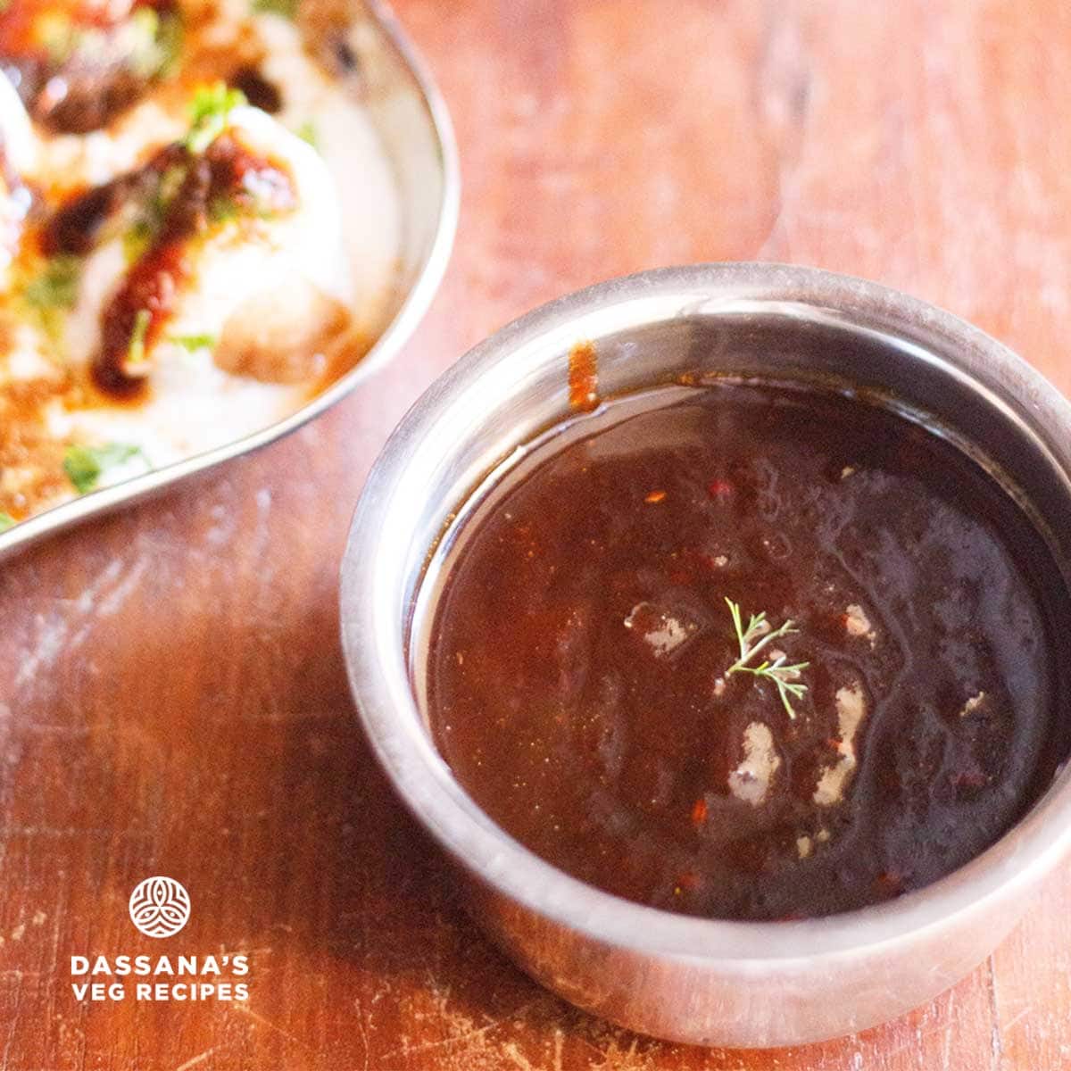 tamarind chutney garnished with one sprig of coriander served in a steel bowl with a square shaped plate of Dahi Vada kept on the left.