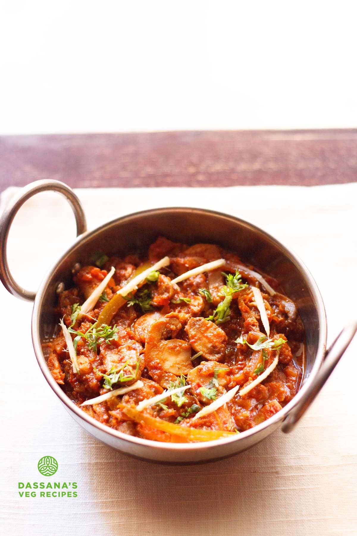 kadai mushroom garnished with coriander leaves, ginger juliennes and served in a steel kadai on a white napkin on a dark ebony colored board.