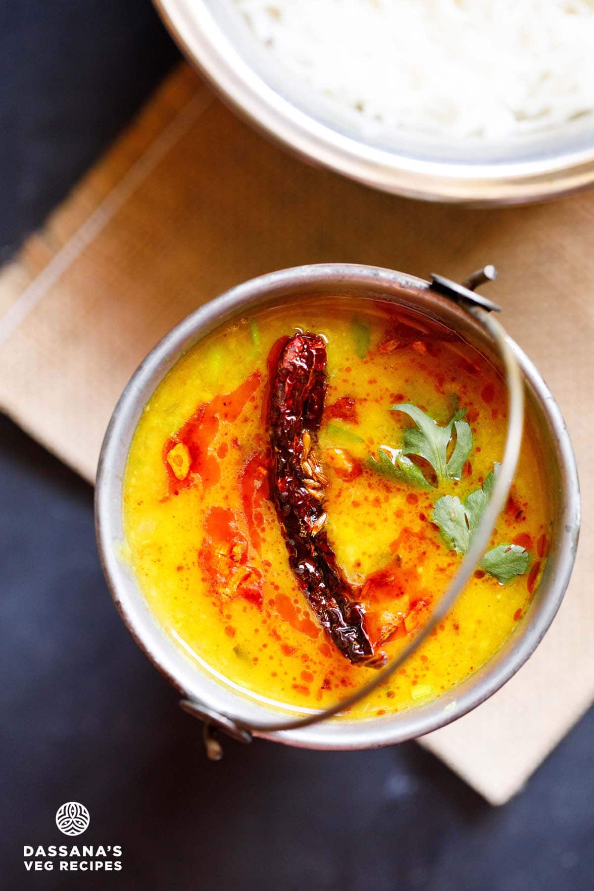 overhead shot of dal tadka garnished with cilantro and a topped with fried red chilli with some fried cumin and some red colored oil in a small brass bucket on a light brown jute mat with a bowl of steamed rice kept above on the top-right.